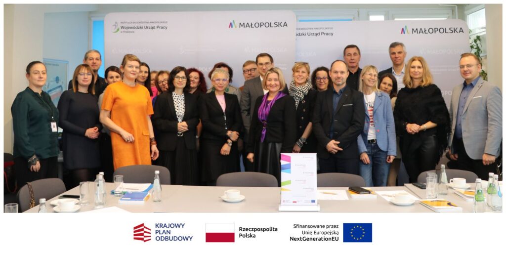 Grupa około 20 dorosłych osób, kobiet i mężczyzn, stoi uśmiechnięta w jasnej sali konferencyjnej. Za nimi znajdują się banery z logo "MAŁOPOLSKA". Na stole przed nimi leżą dokumenty, butelki z wodą i szklanki.