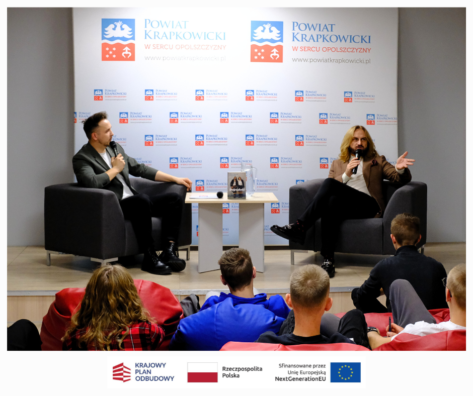 Dwóch mężczyzn siedzi w fotelach na scenie podczas dyskusji, jeden trzyma mikrofon i gestykuluje. Stoją twarzą do publiczności siedzącej na workach z fasolą. Za nimi znajduje się baner Powiatu Krapkowickiego z logo. Logotypy i flagi konferencji pojawiają się w dolnej części obrazu.