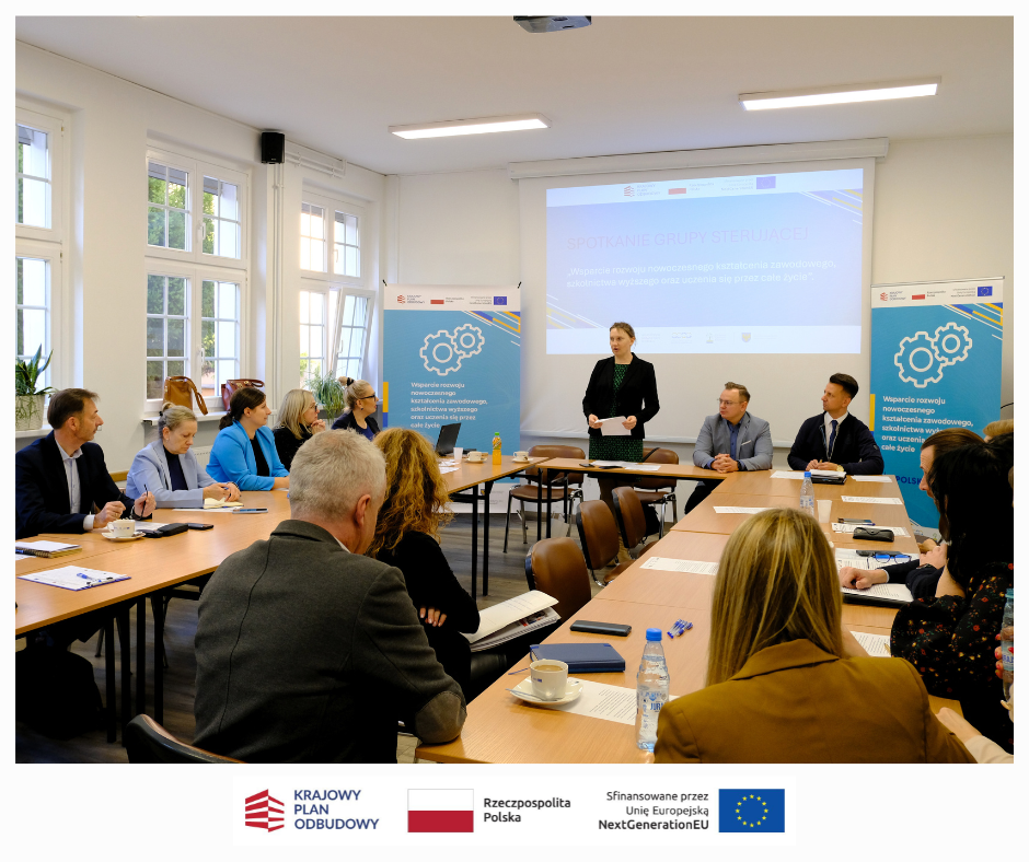 Grupa ludzi siedzi przy prostokątnych stołach w jasnej sali konferencyjnej. Kobieta stoi i przemawia w pobliżu dużego ekranu wyświetlającego prezentację. W tle widoczne są banery z kołami zębatymi i logo, a także okna.
