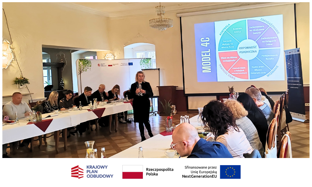 Kobieta stoi i przemawia do grupy siedzącej wokół stołów w kształcie litery U w sali konferencyjnej. Na dużym ekranie wyświetlany jest kolorowy diagram modelu 4C. Uczestnicy słuchają uważnie; notatki, napoje i jedzenie znajdują się na stołach w jasnej, eleganckiej przestrzeni.