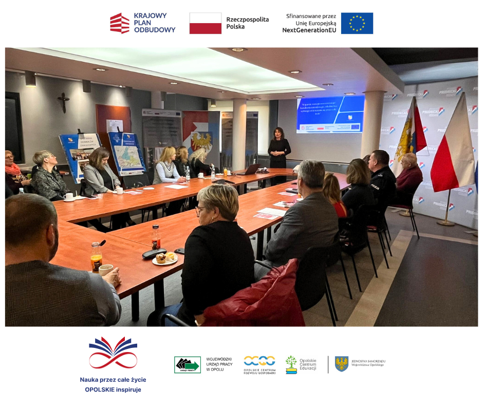Grupa ludzi siedzi wokół stołu konferencyjnego w kształcie litery U, słuchając kobiety prezentującej informacje wyświetlane na ekranie z przodu. Na stole leżą dokumenty, kubki i przekąski. W tle widoczne są flagi i plakaty informacyjne.