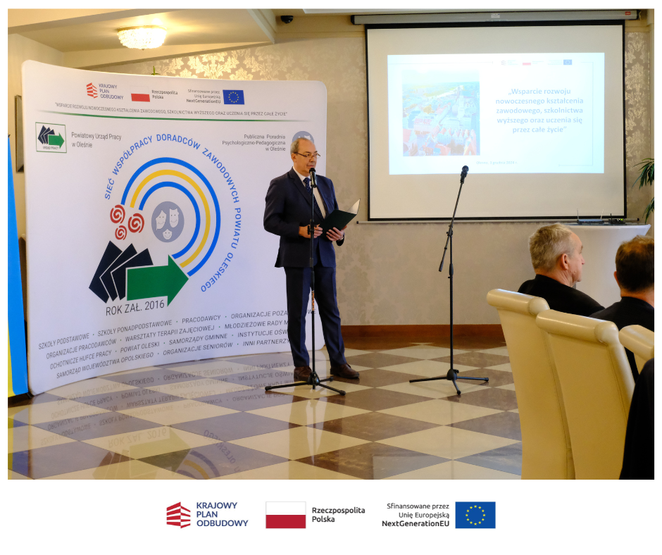 Mężczyzna w garniturze przemawia na podium w sali konferencyjnej, stojąc obok dużego banera z polskim tekstem i logo. Za nim ekran projektora wyświetla slajd. Dwóch siedzących mężczyzn słucha uważnie. Logotypy i informacje o finansowaniu są widoczne u dołu.