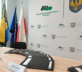 Sala konferencyjna ze stołem, na którym leżą stosy czarnych teczek, szklanka wody, długopis i tabliczka z nazwiskiem Macieja Kalskiego. Z tyłu cztery flagi i tło z logo Wojewódzkiego Urzędu Pracy w Opolu i herbem Opolszczyzny.