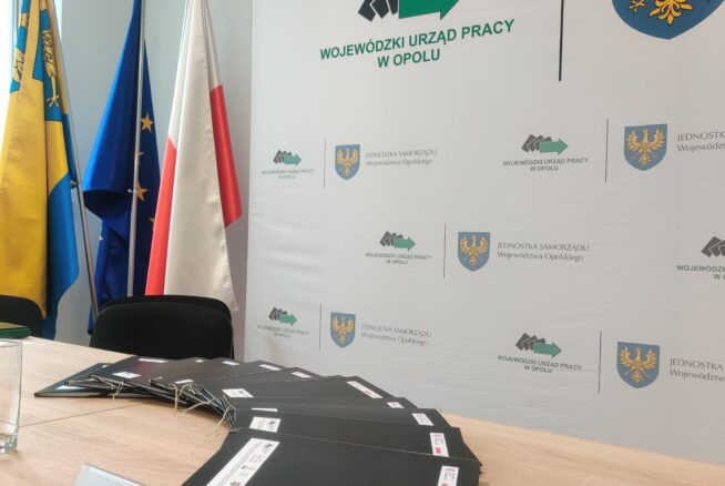 Sala konferencyjna ze stołem, na którym leżą stosy czarnych teczek, szklanka wody, długopis i tabliczka z nazwiskiem Macieja Kalskiego. Z tyłu cztery flagi i tło z logo Wojewódzkiego Urzędu Pracy w Opolu i herbem Opolszczyzny.