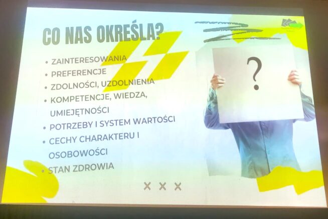 Slajd prezentacji w języku polskim zatytułowany CO NAS OKREŚLA? wymienia cechy, które nas definiują, takie jak zainteresowania, preferencje, zdolności, umiejętności, wiedza, wartości, osobowość i zdrowie. Po prawej stronie osoba trzyma papier z dużym znakiem zapytania zakrywającym jej twarz.