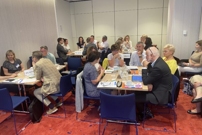 Grupa ludzi siedzi w małych grupach przy stołach w jasno oświetlonej sali konferencyjnej, zaangażowana w dyskusję. Na stołach leżą papiery, notatniki i tabliczki z nazwiskami. Większość osób wydaje się słuchać lub rozmawiać, tworząc aktywną, opartą na współpracy atmosferę.