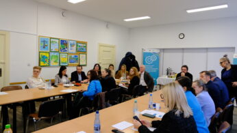Grupa ludzi, mężczyzn i kobiet, siedzi wokół dwóch prostokątnych stołów ustawionych w kształcie litery U w jasnej sali konferencyjnej. Kolorowe rysunki dzieci zdobią białą ścianę. Na stołach znajduje się woda butelkowana, gazety i przekąski. W tle wisi niebieski baner.