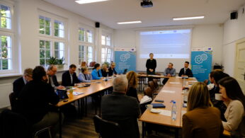 Grupa ludzi siedzi wokół trzech połączonych stołów w jasnej sali konferencyjnej z dużymi oknami. Kobieta stoi z przodu, prezentując grupie. Za nią widoczne są slajdy prezentacji i niebieskie banery z ilustracjami białych kół zębatych.