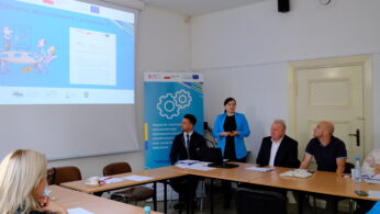 Kobieta stoi i przemawia obok ekranu wyświetlającego prezentację w sali konferencyjnej. Cztery siedzące osoby słuchają uważnie wokół stołu z papierami, butelkami i laptopami. W pobliżu prezenterki stoi niebieski baner z symbolami kół zębatych.
