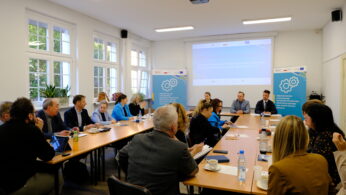 Grupa ludzi siedzi przy prostokątnych stołach w sali konferencyjnej z dużymi oknami. Są zaangażowani w spotkanie, patrząc na prezentację wyświetlaną na ekranie z przodu. Po obu stronach ekranu wyświetlane są dwa niebieskie banery z grafiką kół zębatych.
