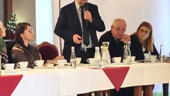 Mężczyzna w garniturze stoi i mówi do mikrofonu przy stole konferencyjnym. Cztery siedzące osoby słuchają, z kubkami, szklankami i butelkami na stole. W tle biały baner z polskim tekstem i logo. Atmosfera jest formalna i uważna.