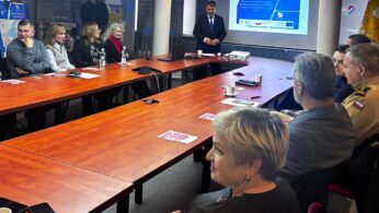 Grupa ludzi siedzi przy dużym stole konferencyjnym, słuchając mężczyzny w garniturze stojącego przy ekranie. Na ekranie wyświetlany jest niebieski slajd prezentacji. Na stole w nowoczesnej sali konferencyjnej leżą papiery, kubki i notatniki.