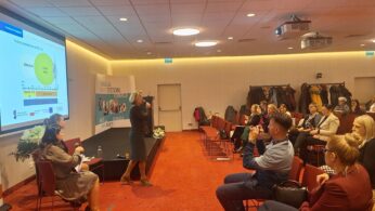 Kobieta stoi na scenie, prezentując slajd z wykresem kołowym publiczności w sali konferencyjnej. Dwie inne kobiety siedzą w pobliżu. Uczestnicy uważnie słuchają; niektórzy robią notatki lub zdjęcia. Płaszcze wiszą na hakach z tyłu. Atmosfera jest profesjonalna i zaangażowana.