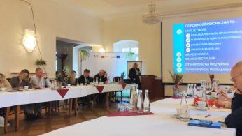 Grupa ludzi siedzi wokół stołów w kształcie litery U w dobrze oświetlonym pomieszczeniu, słuchając mówcy na podium. Na dużym ekranie wyświetlany jest niebieski slajd w języku polskim z ponumerowaną listą. Na stołach leżą butelki, kubki i notatniki.