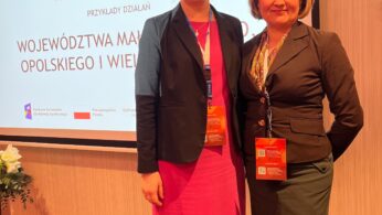 Dwie kobiety stoją obok siebie, uśmiechając się do kamery. Obie mają na sobie stroje biznesowe z plakietkami konferencyjnymi na szyjach. Za nimi znajduje się ekran z polskim tekstem o działaniach regionalnych i funduszach europejskich, a po lewej stronie widoczna jest kompozycja kwiatowa.