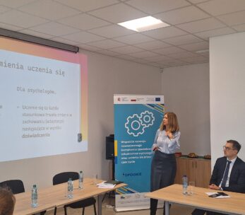 Kobieta stoi i wskazuje na duży ekran projekcyjny z polskim tekstem, przedstawiając się dwóm siedzącym kolegom. Za nią stoi baner. W pokoju znajduje się stół z butelkami wody, notatnikami i sufitem z białymi płytkami i światłami.