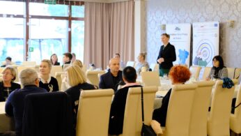 Grupa ludzi siedzi przy okrągłych stołach w jasnej sali konferencyjnej. Jedna z kobiet stoi i zwraca się do uczestników. Za nią znajdują się duże banery z tekstem i grafiką. Atmosfera wydaje się profesjonalna i uważna, a ludzie słuchają i wchodzą w interakcje.