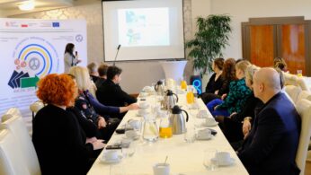 Kobieta stoi z przodu sali konferencyjnej, prezentując slajdy grupie osób siedzących przy długim stole zastawionym napojami i filiżankami. Publiczność słucha uważnie. Na lewej ścianie widoczny jest duży baner z kolorową grafiką i tekstem.