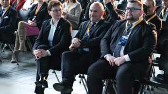 Grupa osób w strojach biznesowych siedzi w rzędach na konferencji. Na pierwszym planie dwóch mężczyzn i jedna kobieta ze smyczami identyfikacyjnymi siedzą uważnie. Wielu innych siedzi za nimi, niektórzy obserwują scenę, w jasno oświetlonym wnętrzu.