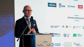 Łysy mężczyzna w garniturze i okularach przemawia na podium z mikrofonem podczas wydarzenia. Za nim na ekranie wyświetlane są logotypy partnerów strategicznych i merytorycznych, w tym mBanku i Uniwersytetu Opolskiego, a na podium logo Opolskiego Centrum Rozwoju Gospodarki.