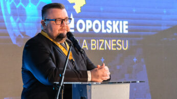 Mężczyzna w garniturze i okularach przemawia na podium z mikrofonem. Za nim znajduje się niebieski ekran wyświetlający żółtą mapę, sześciokątne wzory i napis OPOLSKIE DLA BIZNESU. Na podium znajduje się logo Opolskiego Centrum Rozwoju Gospodarki.