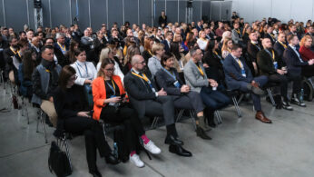 Duża grupa ludzi w strojach biznesowych siedzi w rzędach na konferencji w pomieszczeniu, wielu z nich nosi żółte smycze. Publiczność wydaje się być uważna, niektórzy patrzą przed siebie, inni zerkają na urządzenia. Pomieszczenie ma szare ściany działowe i betonową podłogę.
