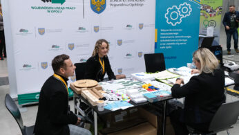 Trzy osoby siedzą przy stoisku wystawowym pokrytym dokumentami, broszurami i kolorowymi plastikowymi modelami. Za nimi znajdują się banery polskich regionalnych urzędów pracy i edukacji. Wygląda to na targi pracy lub edukacji w dużym pomieszczeniu.