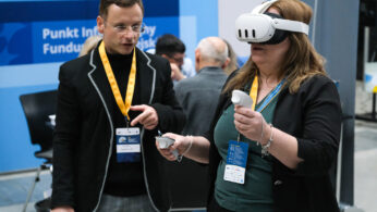 Kobieta nosząca gogle VR i trzymająca dwa kontrolery wchodzi w interakcję z wirtualną zawartością, podczas gdy mężczyzna obok niej gestykuluje i mówi. Oboje noszą smycze z identyfikatorami. W tle widoczne są inne osoby i niebieski znak z polskim tekstem.