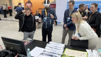 Grupa ludzi, niektórzy noszą smycze, gromadzi się wokół stołu na imprezie wewnętrznej. Jedna osoba korzysta z zestawu VR, podczas gdy inni obserwują ją z zainteresowaniem. Na stole znajdują się broszury, identyfikatory i butelka wody. W tle widoczne są stoiska i banery.