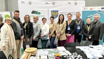 Grupa dwunastu dorosłych osób, mężczyzn i kobiet, stoi w pomieszczeniu przed stoiskami informacyjnymi z banerami i logo. Mają na sobie eleganckie stroje i identyfikatory konferencyjne. Stół na pierwszym planie wyświetla broszury, gadżety i materiały marki.