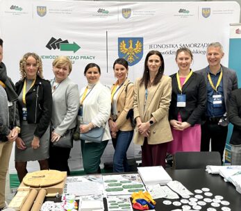 Grupa dwunastu dorosłych osób, mężczyzn i kobiet, stoi w pomieszczeniu przed stoiskami informacyjnymi z banerami i logo. Mają na sobie eleganckie stroje i identyfikatory konferencyjne. Stół na pierwszym planie wyświetla broszury, gadżety i materiały marki.