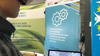 Osoba nosząca gogle VR i trzymająca kontroler stoi w pobliżu monitora komputerowego wyświetlającego wirtualne środowisko. Za nimi znajdują się niebieskie i zielone banery z polskim tekstem o programach rozwoju zawodowego i edukacji technicznej.