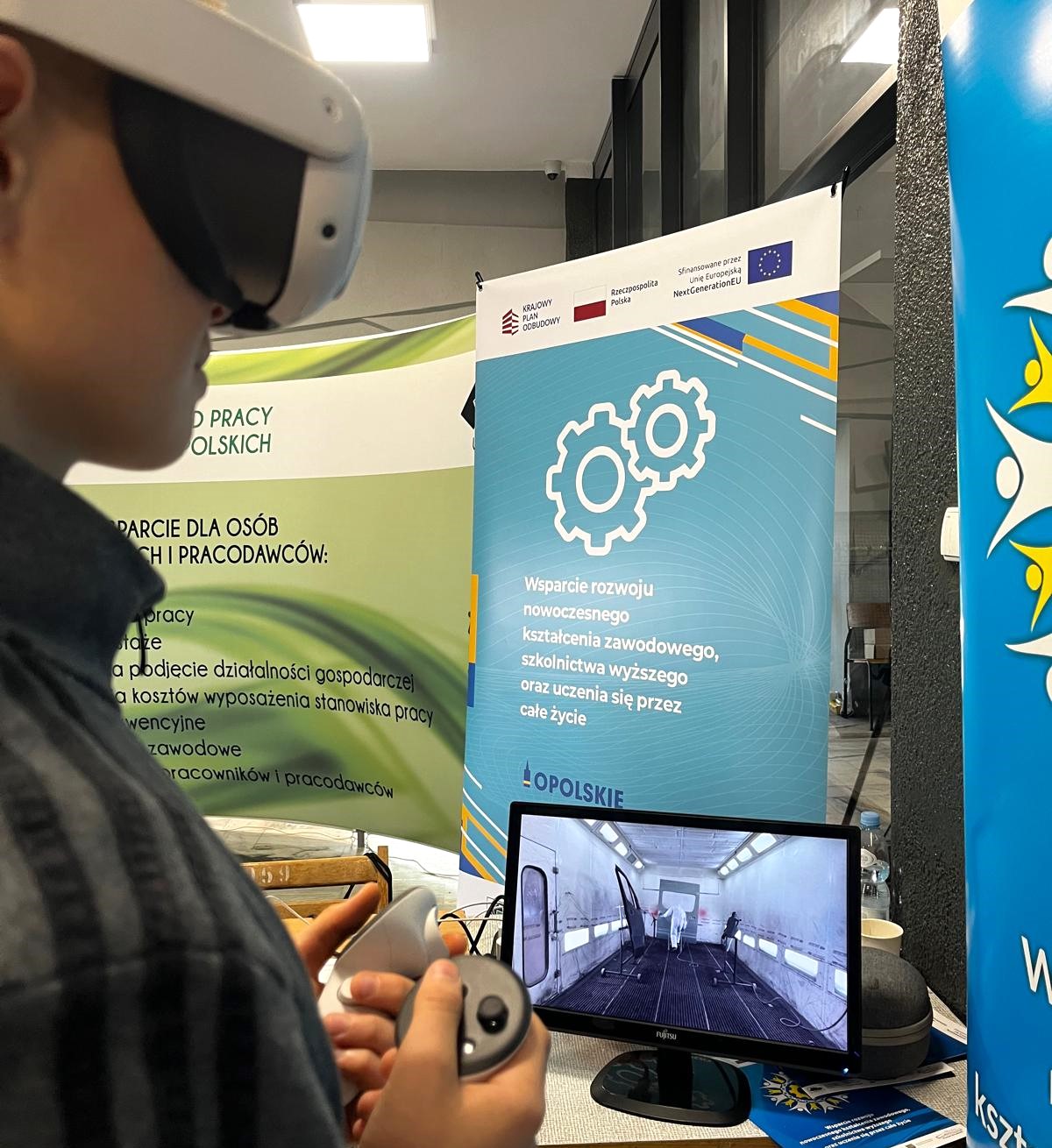 Osoba nosząca gogle VR i trzymająca kontroler stoi w pobliżu monitora komputerowego wyświetlającego wirtualne środowisko. Za nimi znajdują się niebieskie i zielone banery z polskim tekstem o programach rozwoju zawodowego i edukacji technicznej.