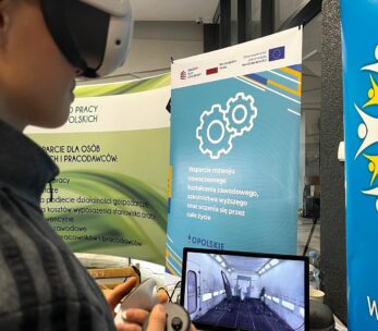 Osoba nosząca gogle VR i trzymająca kontroler wchodzi w interakcję z wirtualnym środowiskiem wyświetlanym na monitorze. Za nimi znajdują się banery promujące nowoczesne szkolenia zawodowe i programy edukacyjne, zawierające logo i symbole sprzętu.