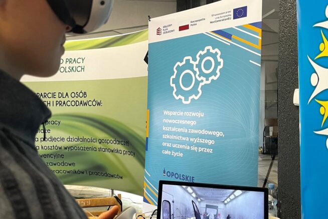 Osoba nosząca gogle VR i trzymająca kontroler wchodzi w interakcję z wirtualnym środowiskiem wyświetlanym na monitorze. Za nimi znajdują się banery promujące nowoczesne szkolenia zawodowe i programy edukacyjne, zawierające logo i symbole sprzętu.