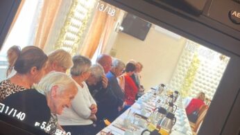 Kamera pokazuje grupę starszych osób siedzących przy długim stole zastawionym białymi obrusami, naczyniami i dzbanami. Wydaje się, że słuchają uważnie w jasnym pokoju z dużymi oknami i wzorzystymi zasłonami.