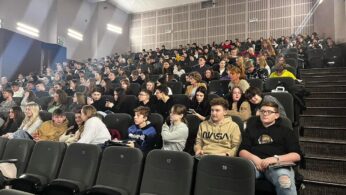 Duża grupa, głównie młodych ludzi, siedzi w rzędach ciemnych foteli w nowoczesnym audytorium. Pomieszczenie jest dobrze oświetlone, a niektóre miejsca w pierwszym rzędzie są puste. Większość uczestników ma na sobie luźne ubrania i wydaje się uważna lub zaangażowana.