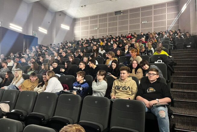 Duża grupa, głównie młodych ludzi, siedzi w rzędach ciemnych foteli w nowoczesnym audytorium. Pomieszczenie jest dobrze oświetlone, a niektóre miejsca w pierwszym rzędzie są puste. Większość uczestników ma na sobie luźne ubrania i wydaje się uważna lub zaangażowana.