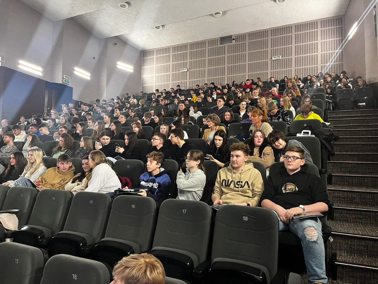 Duża grupa, głównie młodych ludzi, siedzi w rzędach ciemnych foteli w nowoczesnym audytorium. Pomieszczenie jest dobrze oświetlone, a niektóre miejsca w pierwszym rzędzie są puste. Większość uczestników ma na sobie luźne ubrania i wydaje się uważna lub zaangażowana.