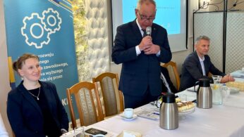 Trzy osoby siedzą przy stole konferencyjnym z mikrofonami, notatnikami, kawą i ciastkami. Mężczyzna w garniturze stoi i mówi do mikrofonu. Widoczny jest baner z ikonami kół zębatych i polskim tekstem oraz ekran pokazujący niebieski slajd prezentacji w tle.