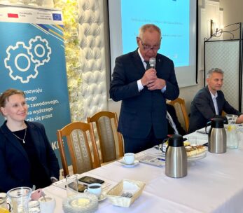 Trzy osoby siedzą przy stole konferencyjnym z mikrofonami, notatnikami, kawą i ciastkami. Mężczyzna w garniturze stoi i mówi do mikrofonu. Widoczny jest baner z ikonami kół zębatych i polskim tekstem oraz ekran pokazujący niebieski slajd prezentacji w tle.