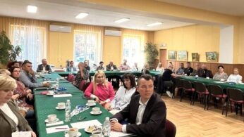 Grupa dorosłych siedzi wokół długich zielonych stołów w sali konferencyjnej z żółtymi ścianami, dużymi oknami i roślinami doniczkowymi. Wiele osób stoi twarzą do kamery, niektórzy z notatnikami i napojami. Na ścianach wiszą obrazy, a atmosfera wydaje się formalna i uważna.