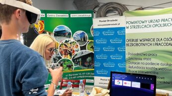 Młody mężczyzna noszący zestaw VR trzyma kontrolery, podczas gdy kobieta siedzi przy stole z materiałami informacyjnymi. Za nimi znajdują się banery w języku polskim promujące usługi samorządowe i wsparcie zatrudnienia. Monitor na stole wyświetla wirtualny interfejs.