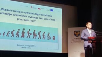 Mężczyzna w garniturze przemawia z podium na scenie obok dużego ekranu. Na ekranie wyświetlany jest polski tekst o uczeniu się przez całe życie, z obrazami ludzi starzejących się od niemowlęcia do osoby starszej. Herb i więcej tekstu są widoczne na znaku obok podium.