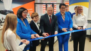 Grupa siedmiu osób, w tym cztery kobiety w niebieskich garniturach, jeden mężczyzna w czarnym garniturze przecinający nożyczkami niebieską wstęgę i dwie kobiety w białych bluzkach, stoją w pomieszczeniu podczas ceremonii przecinania wstęgi. W tle widoczne są maszyny i kolorowe banery.