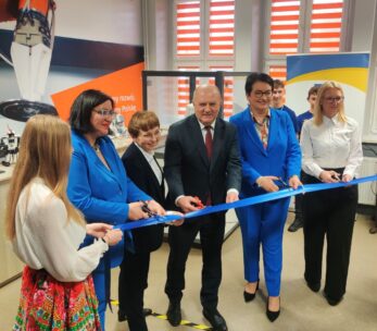 Grupa siedmiu osób, w tym cztery kobiety w niebieskich garniturach, jeden mężczyzna w czarnym garniturze przecinający nożyczkami niebieską wstęgę i dwie kobiety w białych bluzkach, stoją w pomieszczeniu podczas ceremonii przecinania wstęgi. W tle widoczne są maszyny i kolorowe banery.