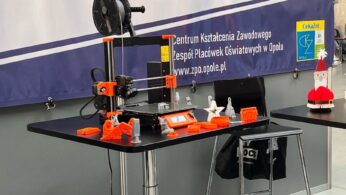 Drukarka 3D stoi na czarnym stole, otoczona małymi pomarańczowymi i białymi wydrukowanymi obiektami. Za nią wyświetlany jest baner z polskim tekstem i obrazami rąk pracujących z narzędziami. Obok znajduje się kolejny pusty czarny stół.