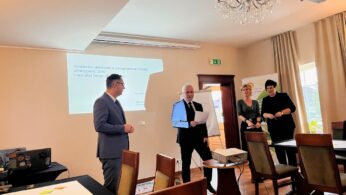 Cztery dorosłe osoby stoją w dobrze oświetlonej sali konferencyjnej z żyrandolem i projektorem. Jeden mężczyzna w garniturze trzyma gazetę, zwracając się do grupy; pozostali słuchają, uśmiechając się. Na ścianie wyświetlana jest prezentacja, a na stołach leżą papiery i długopisy.