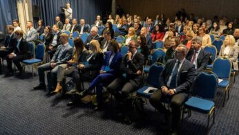 Duża grupa ludzi w strojach biznesowych siedzi w rzędach niebieskich krzeseł na konferencji lub seminarium w sali wyłożonej wykładziną. Większość uczestników jest uważnie zwrócona do przodu. Fotograf stoi z tyłu, a niebieskie zasłony wzdłuż lewej ściany.