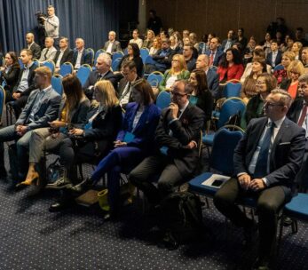 Duża grupa ludzi w strojach biznesowych siedzi w rzędach niebieskich krzeseł na konferencji lub seminarium w sali wyłożonej wykładziną. Większość uczestników jest uważnie zwrócona do przodu. Fotograf stoi z tyłu, a niebieskie zasłony wzdłuż lewej ściany.